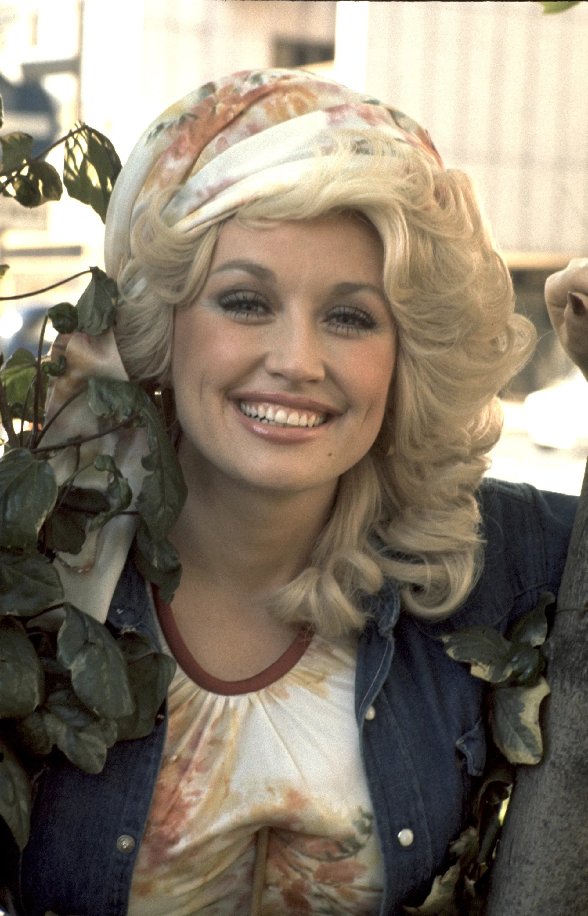Dolly Parton smiling, wearing a headscarf, a printed shirt, and a denim jacket.