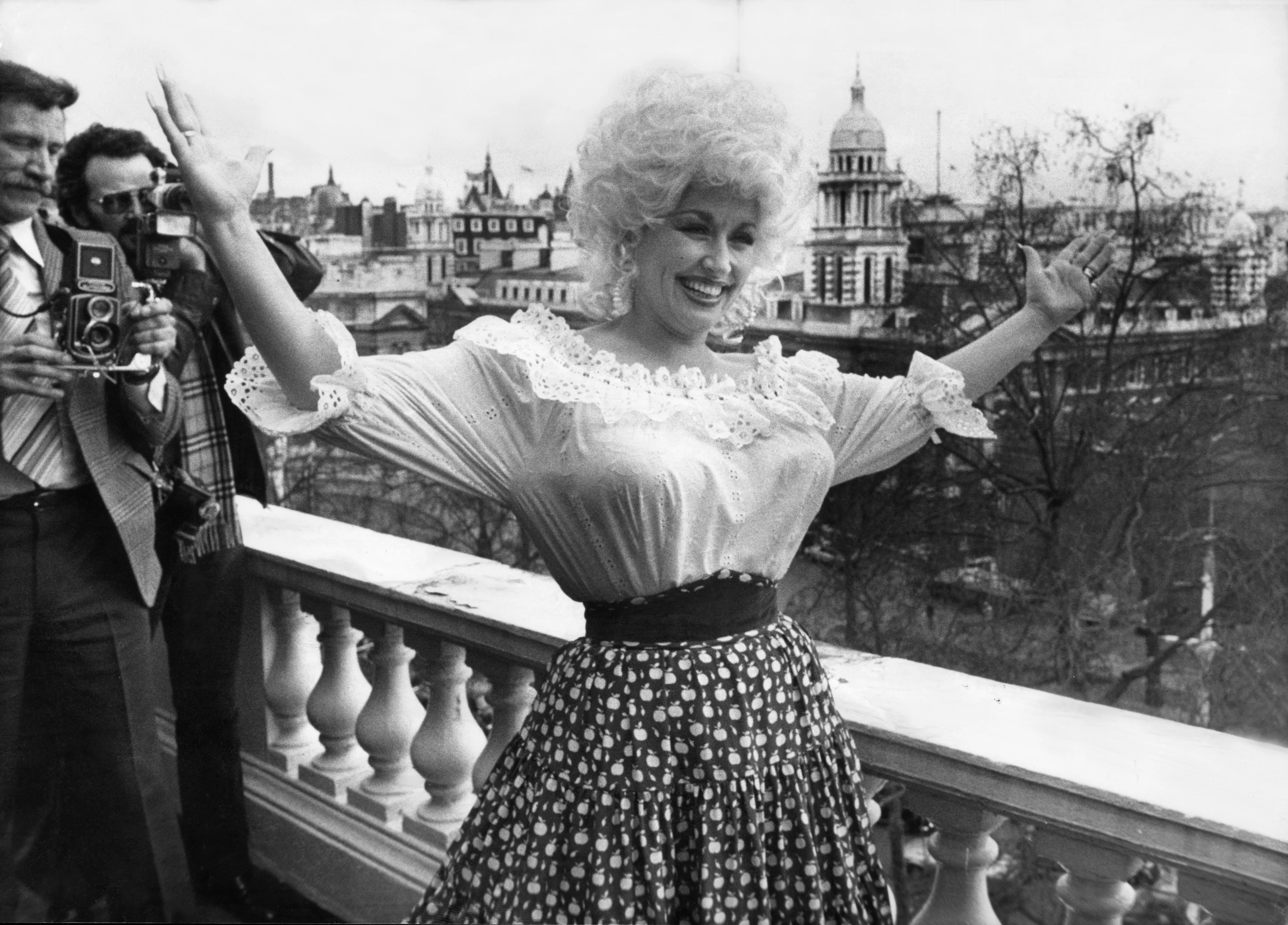 Dolly Parton smiling with her arms outstretched on a balcony overlooking London.