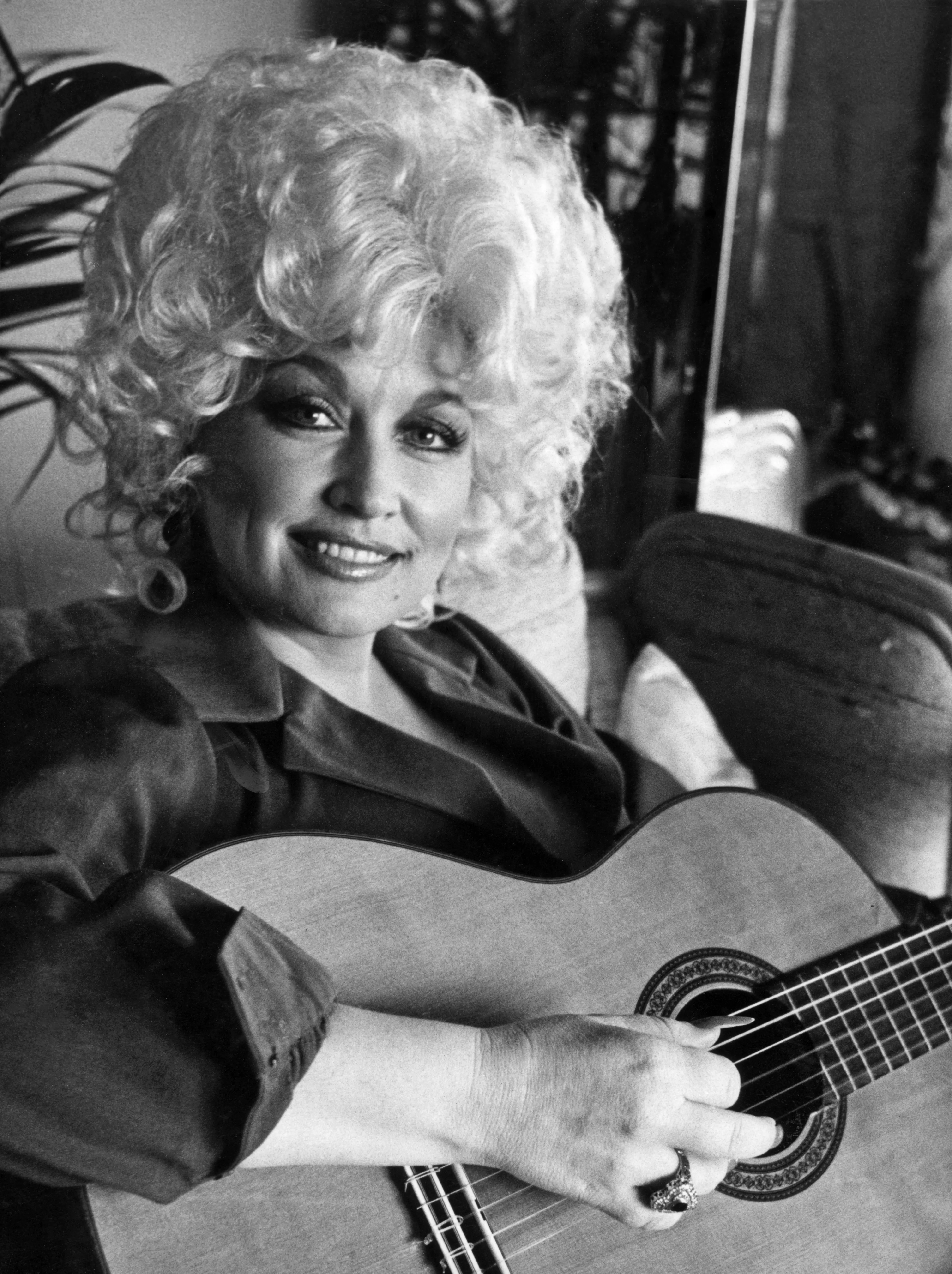 Black and white photo of Dolly Parton, with her blonde hair, smiling at the camera while holding a guitar.