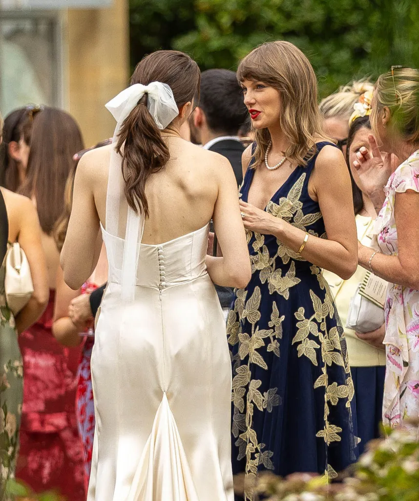 Taylor Swift (right), wearing a navy dress with gold embroidery and a pearl necklace, talking to a guest in a white gown with her back to the camera.