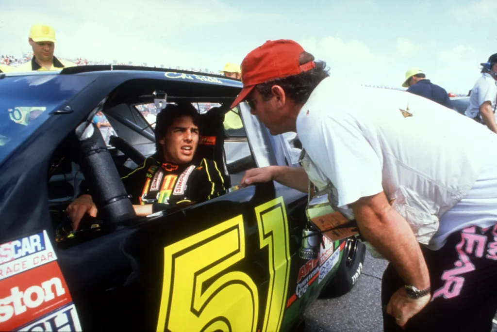 Tom Cruise in a race car talking to a man in a red hat at a track.