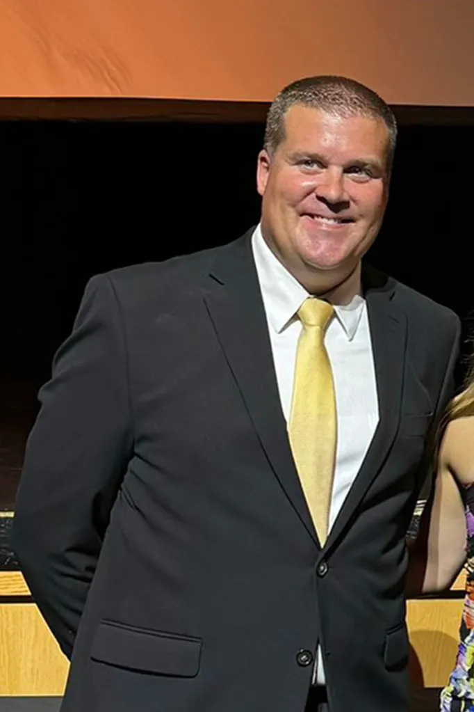 A man in a black suit with a gold tie smiles.