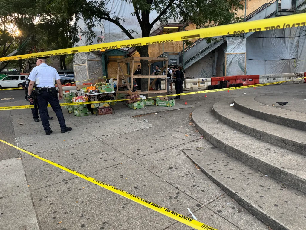 Police investigate a crime scene on a sidewalk corner, marked off with yellow tape, where an overturned fruit stand sits near a subway entrance.
