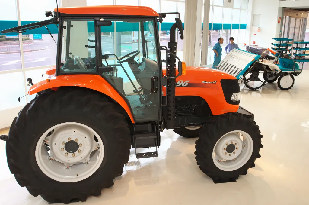 An orange Kubota Corp. tractor on display in a showroom, with two men and other farming equipment in the background.