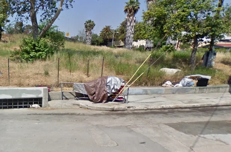 A tarp-covered bed frame and trash on a curb next to a grassy hill.