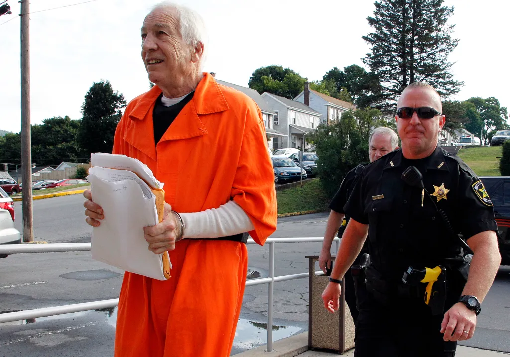 Jerry Sandusky, wearing an orange jumpsuit and handcuffs, escorted by two police officers, enters the Centre County Courthouse.