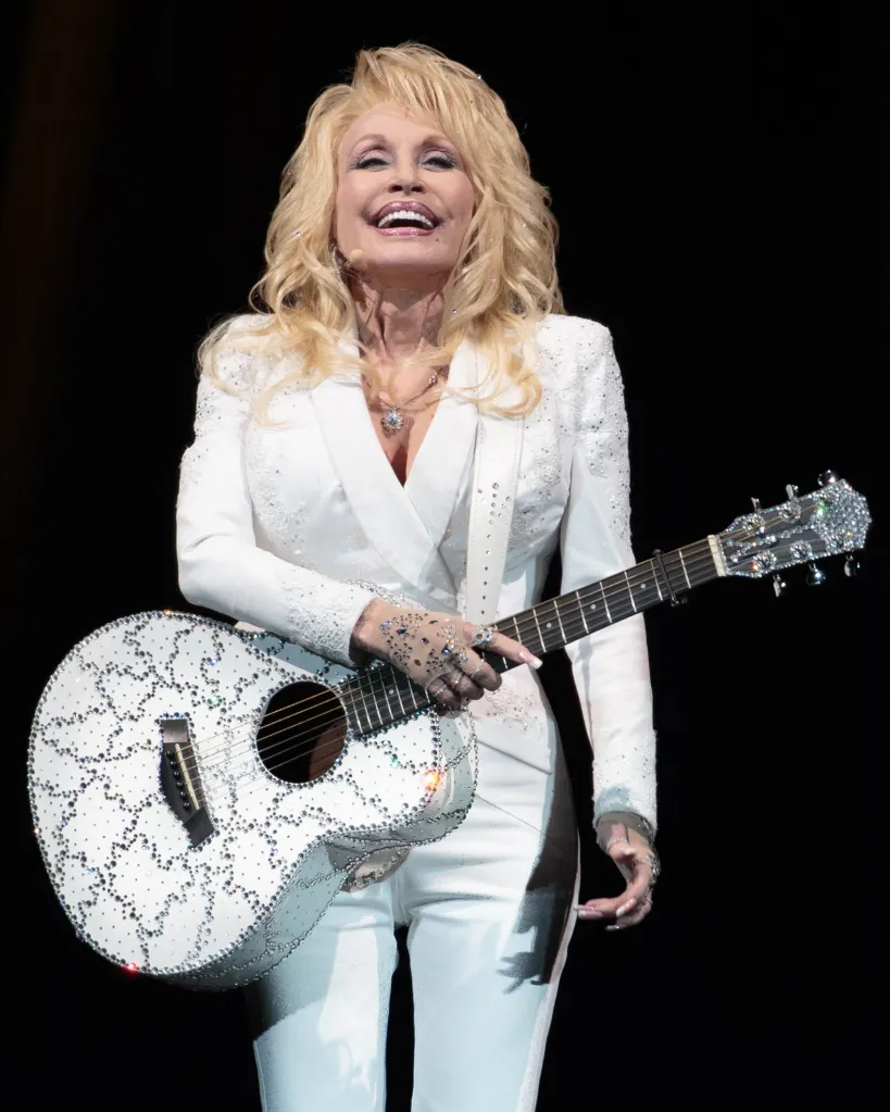Dolly Parton performs in concert, holding a sparkling white guitar and wearing a white pantsuit.