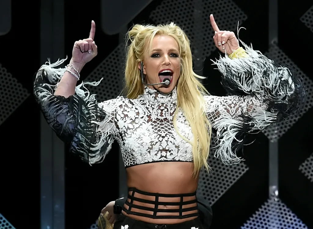 Britney Spears performing with her hands raised, wearing a white lace crop top with feathery sleeves.
