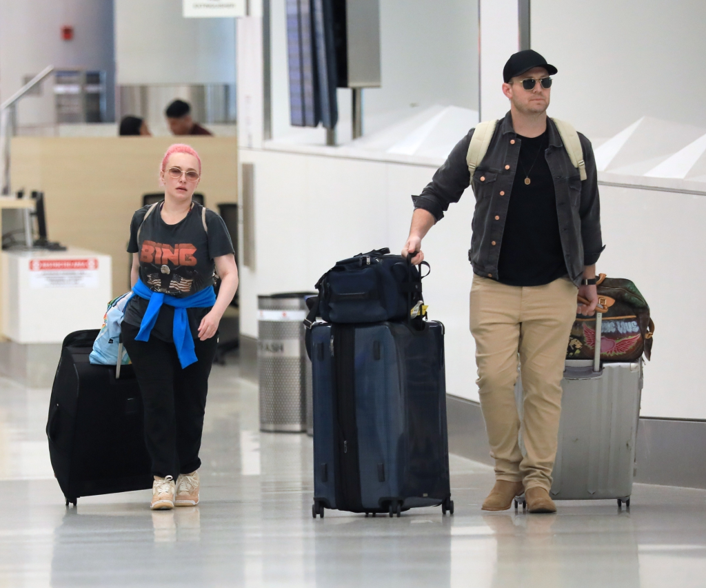 Hayden Panettiere with pink hair and sunglasses pulling a large black suitcase, and Brian Hickerson in a black shirt, khaki pants, and sunglasses pulling two suitcases at LAX airport.