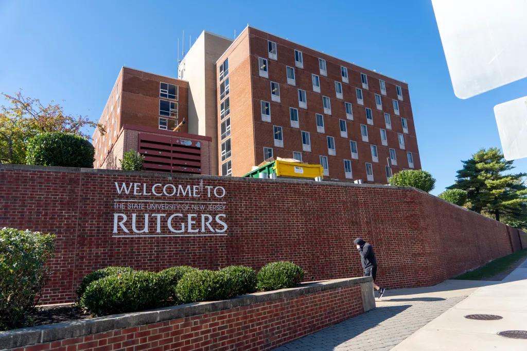 A Rutgers University welcome sign on a brick wall, with dormitories and a person in a hooded sweatshirt nearby.