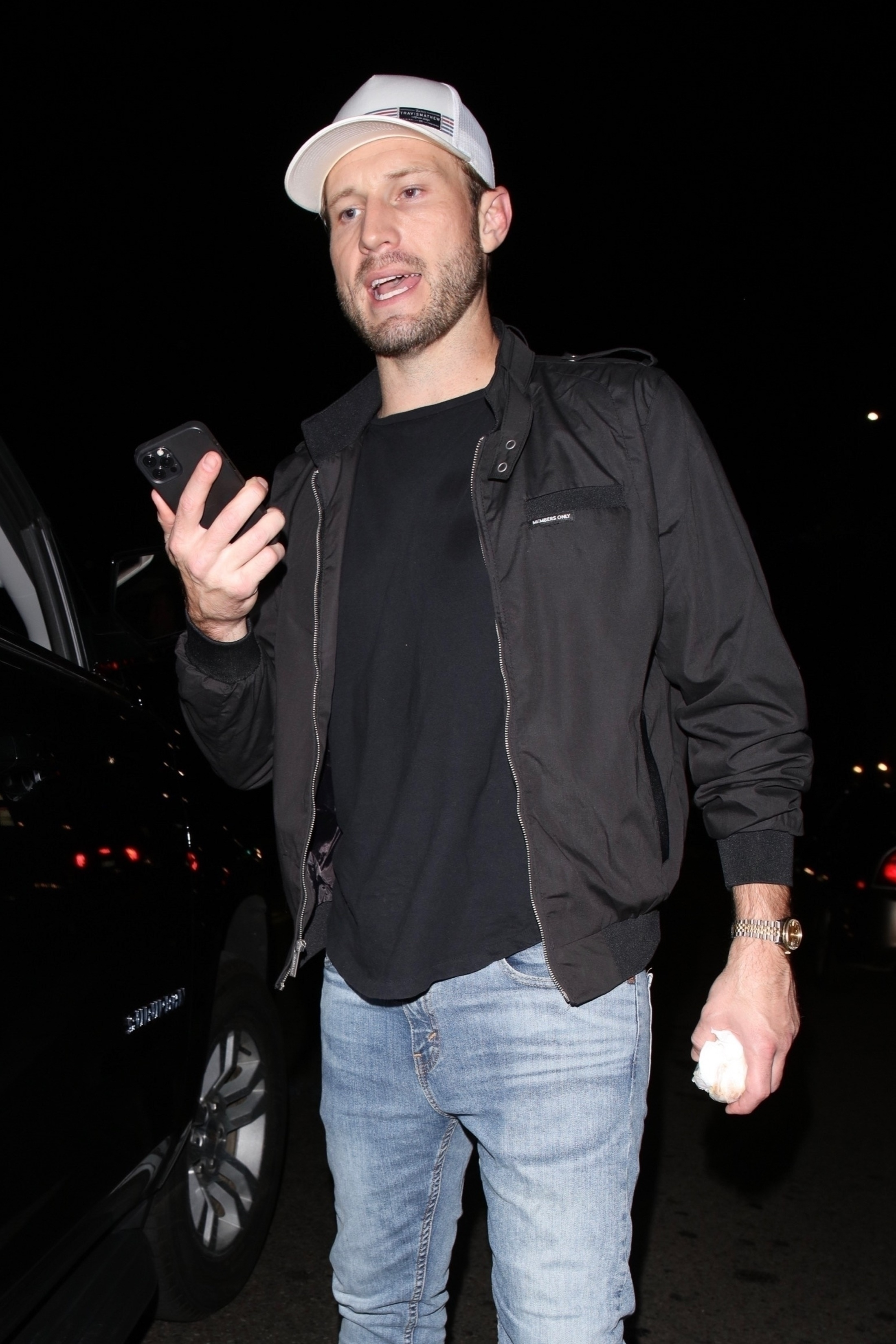 Brian Hickerson wearing a white hat, black jacket, and blue jeans, holding a phone and a crumpled white item.