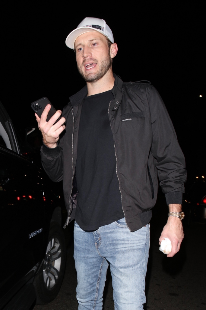 Brian Hickerson wearing a white hat, black jacket, and jeans while holding a phone and a crumpled white item.