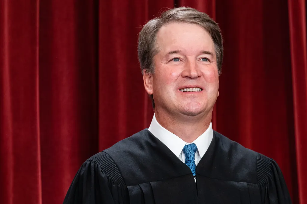 Associate Justice Brett Kavanaugh smiling against a red curtain background.