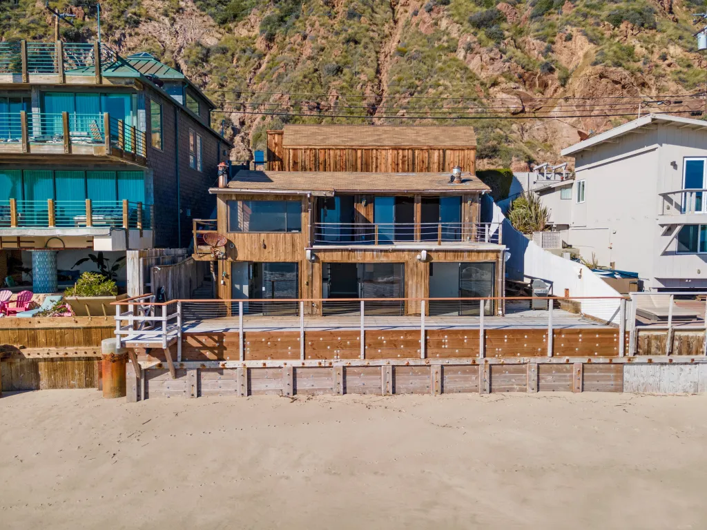 A multi-story beach house with a wooden facade and a deck stands on a sandy beach, with a rocky, vegetated hillside behind it.