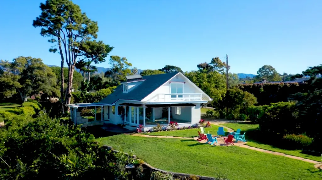 A white house with a dark roof and a grassy yard with colorful chairs.