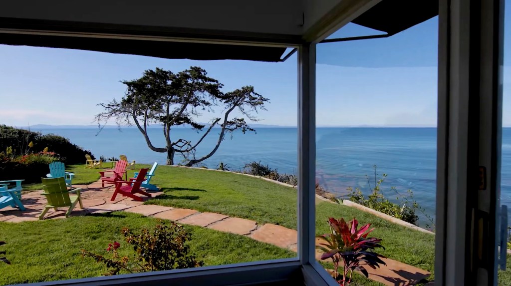 A view of the ocean from a grassy property with colorful Adirondack chairs and a large tree.