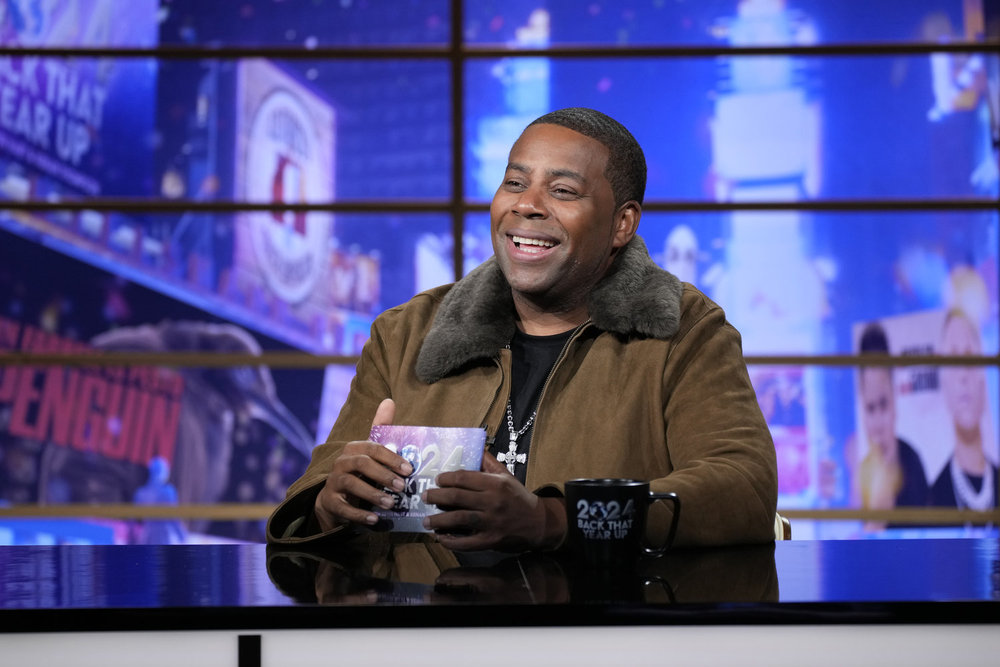 Kenan Thompson smiling while holding a card and sitting at a desk with a mug.