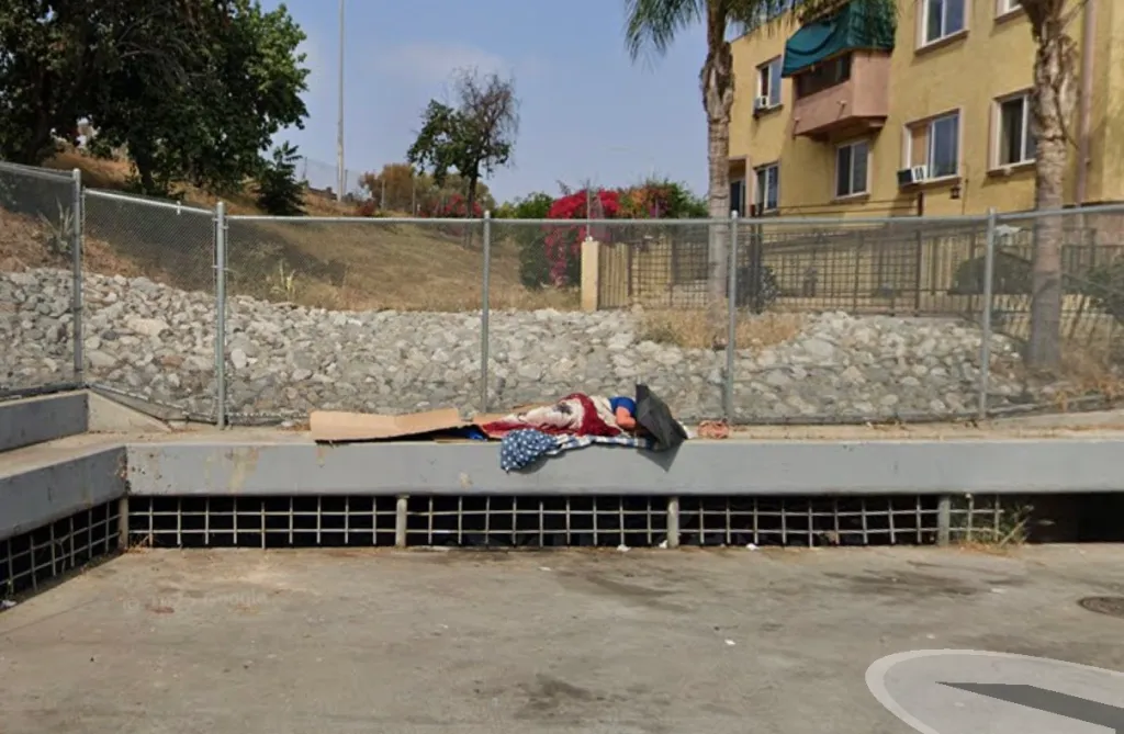 A person sleeping on cardboard and blankets on a ledge next to a chain-link fence.