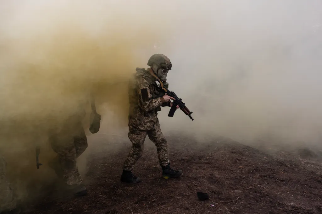 Ukrainian soldier in camouflage, wearing a gas mask and helmet, holding a rifle, surrounded by yellow smoke during military training.