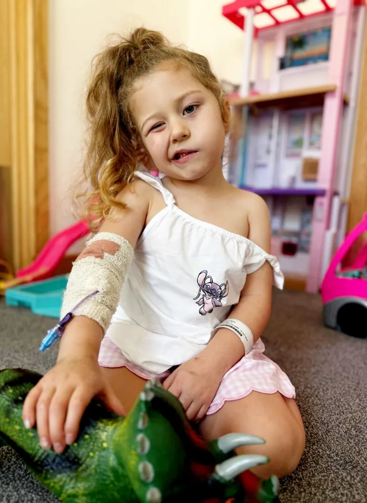 Olivia Elliot, a young girl who suffered a stroke triggered by chickenpox, sits on the floor wearing a hospital bracelet and an IV in her arm.
