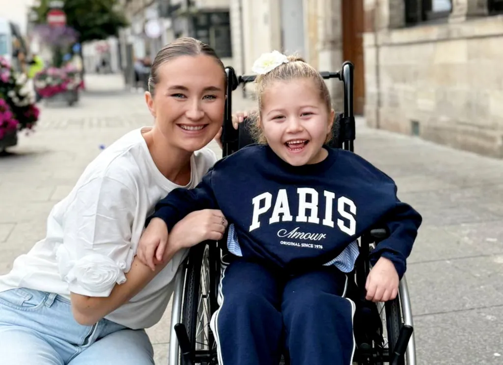 Rebecca Elliot smiles next to her daughter Olivia, who is seated in a wheelchair and also smiling.