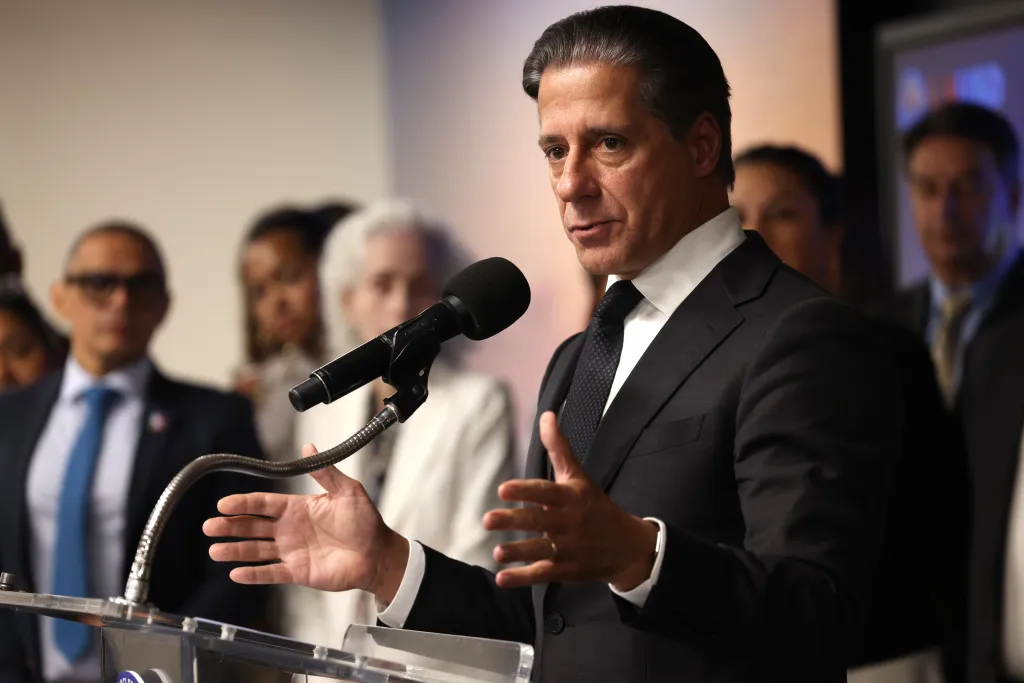 Superintendent Alberto Carvalho speaking at a podium with a microphone, gesturing with his hands, with other people blurred in the background.