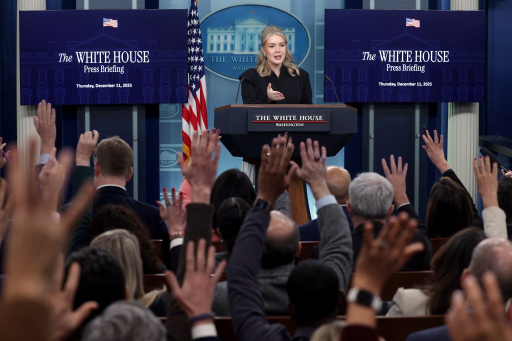 White House Press Secretary Karoline Leavitt speaks from a podium with numerous hands raised by reporters in the foreground.