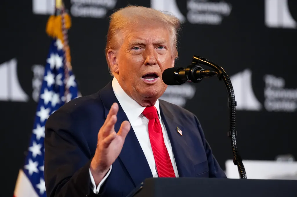 Donald Trump speaking at a podium with a microphone, gesturing with his hand, with an American flag in the background.