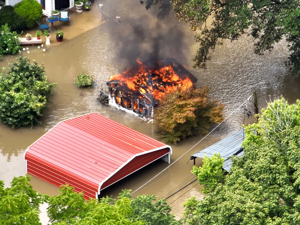 A house on fire, engulfed in flames and smoke, stands in the middle of a flooded street next to another building with a red roof.