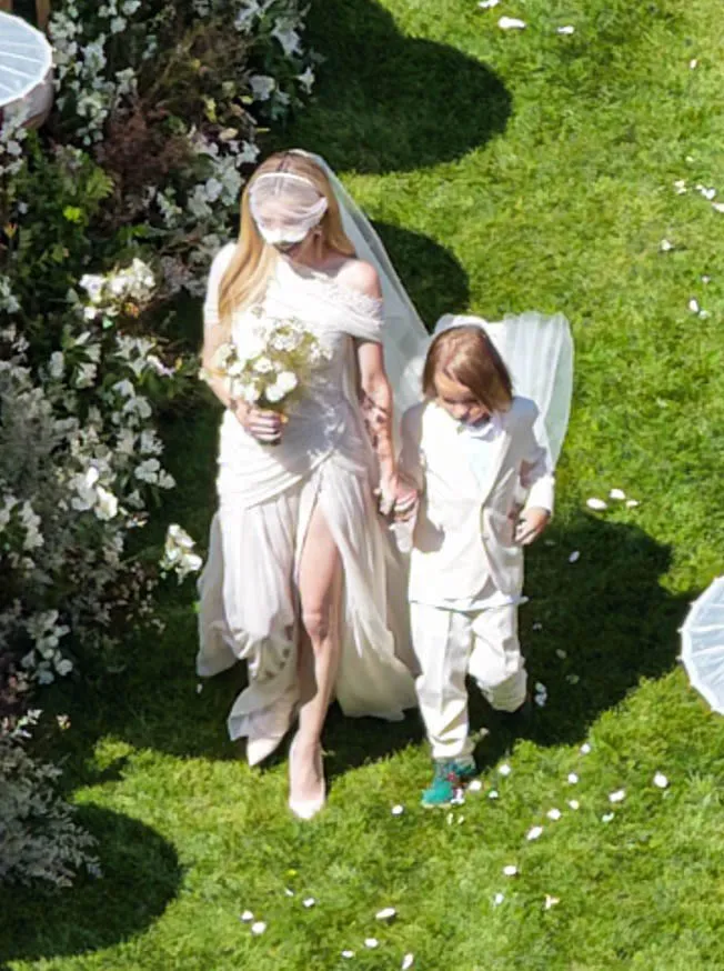 Emma Roberts, wearing a wedding gown and veil, walks with a child in a white suit, holding hands, down a green grassy aisle with white petals scattered around.