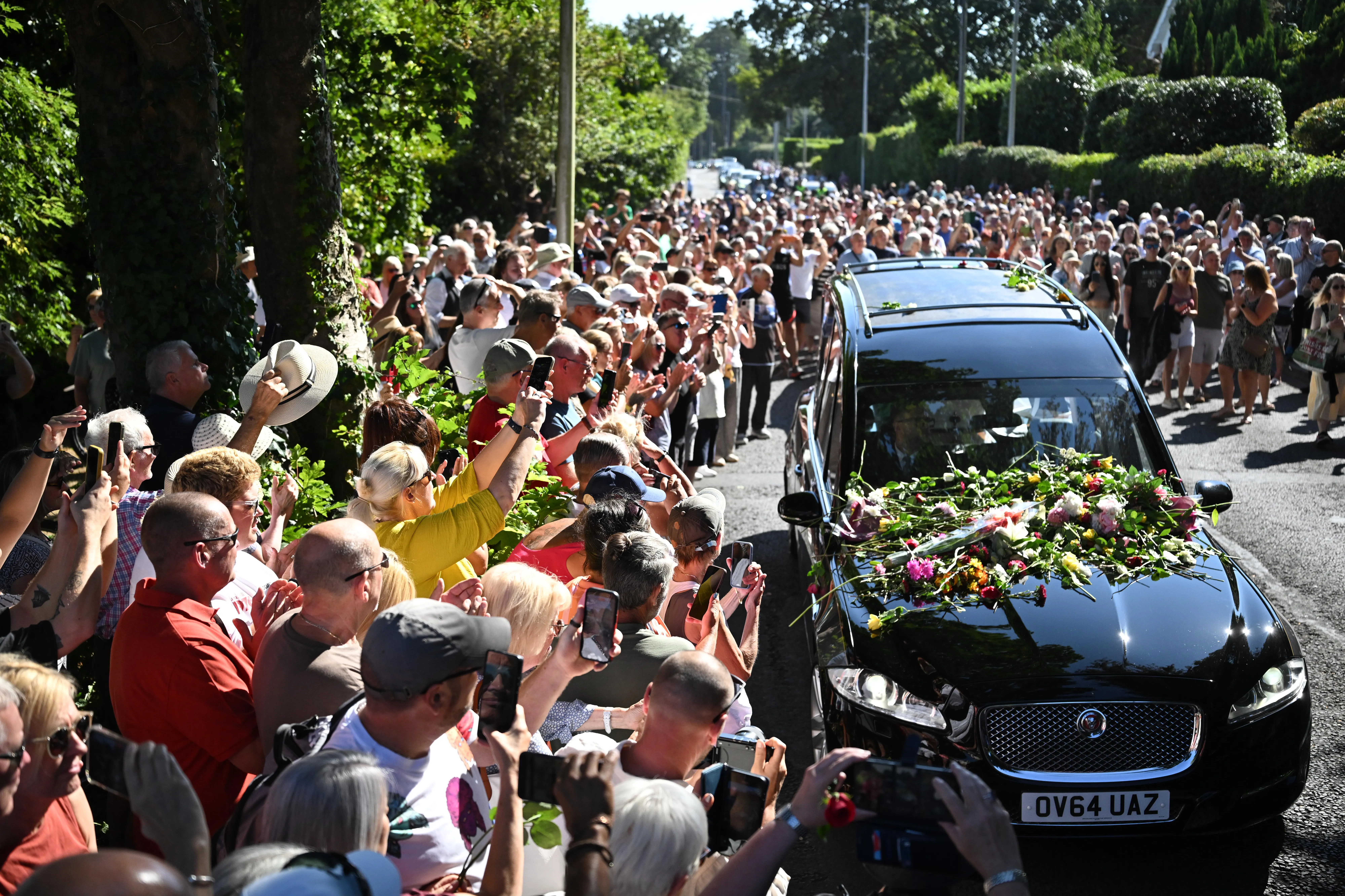 Funeral cortege for Bonnie Tyler, adorned with flowers, passes through a crowd of people.