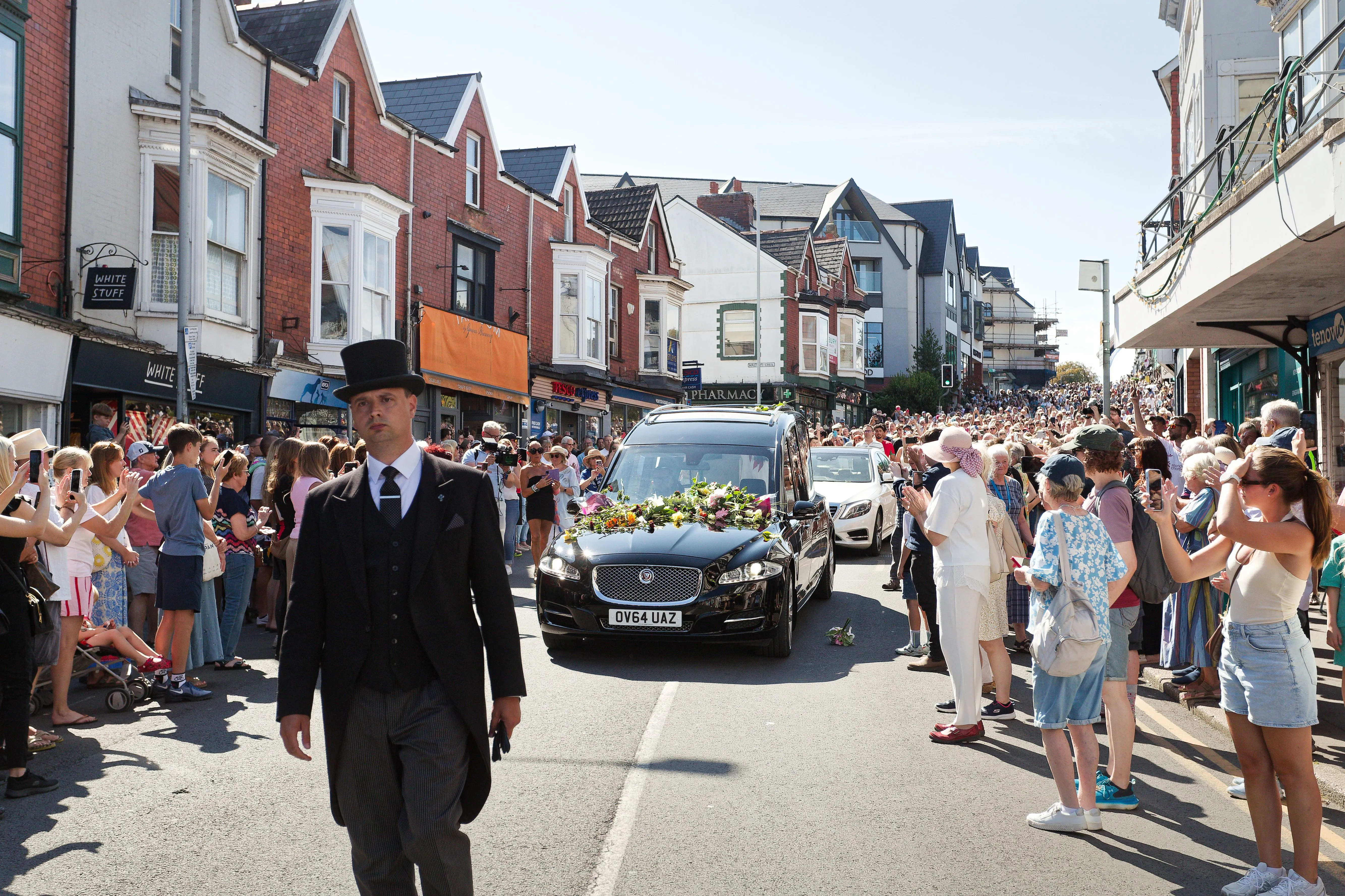 Mourners line the streets as the hearse carrying Bonnie Tyler proceeds down a street lined with buildings and shops.