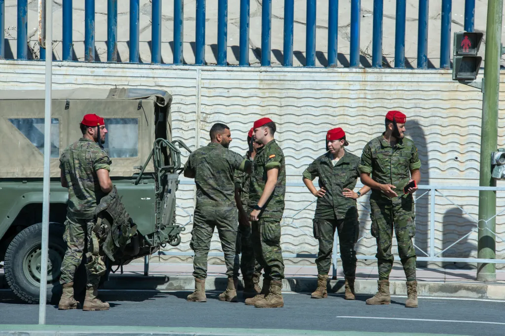 Military personnel in camouflage uniforms and red berets stand near a military vehicle.