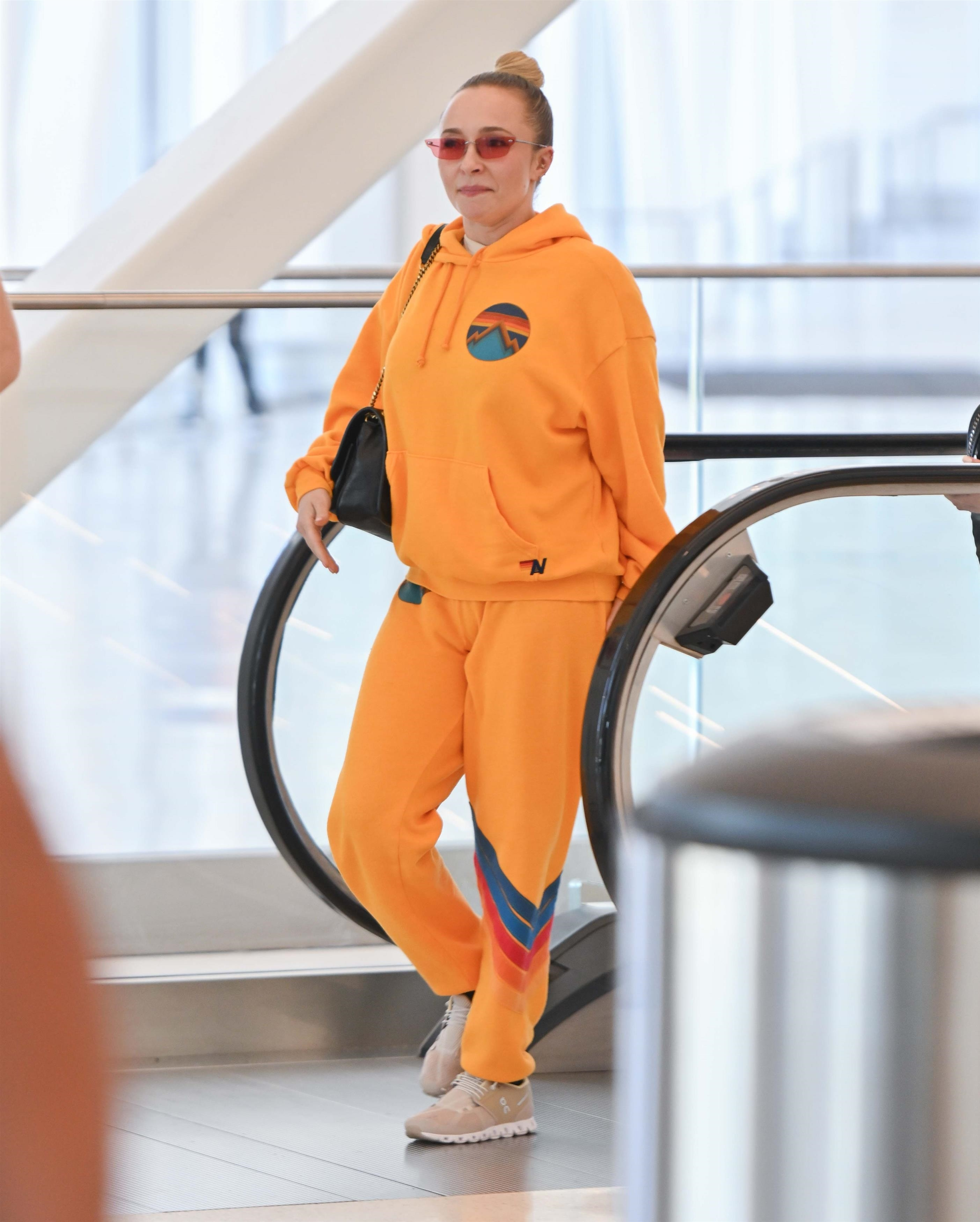 Hayden Panettiere in a bright orange sweatsuit and pink sunglasses, walking through an airport.