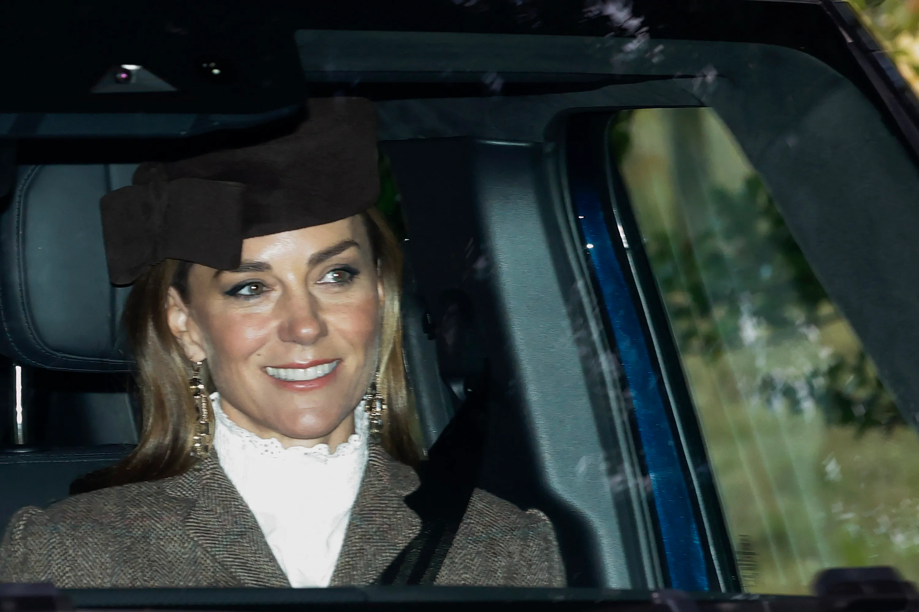 Catherine, Princess of Wales, in a car, smiling while wearing a brown hat, brown tweed jacket, and white ruffled collar.