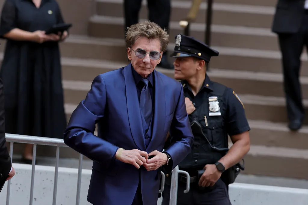 Singer Barry Manilow in a blue suit and sunglasses, walking past a police officer.