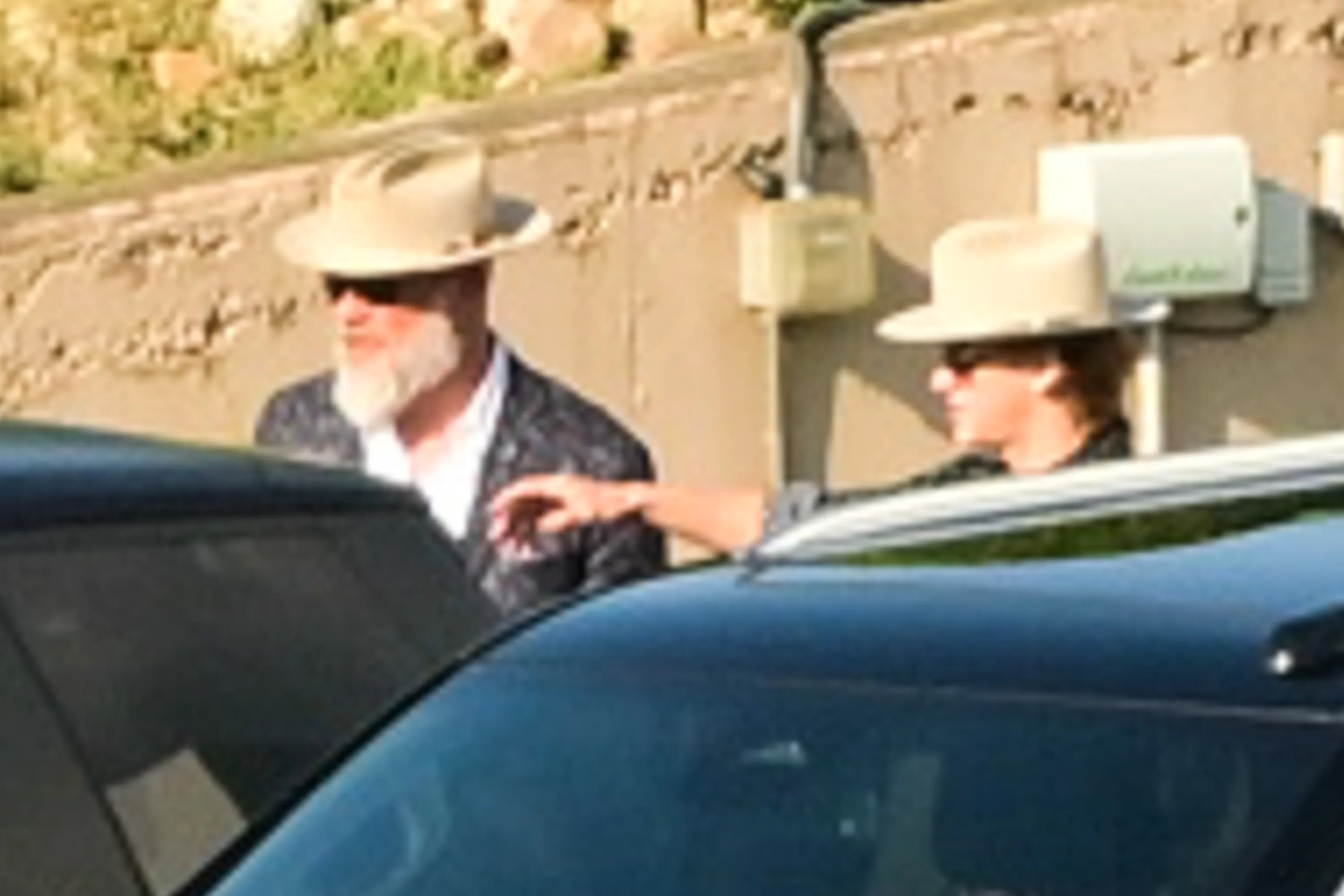 A man in a hat and beard stands by a car, looking away, while another person in a hat reaches an arm forward behind him.