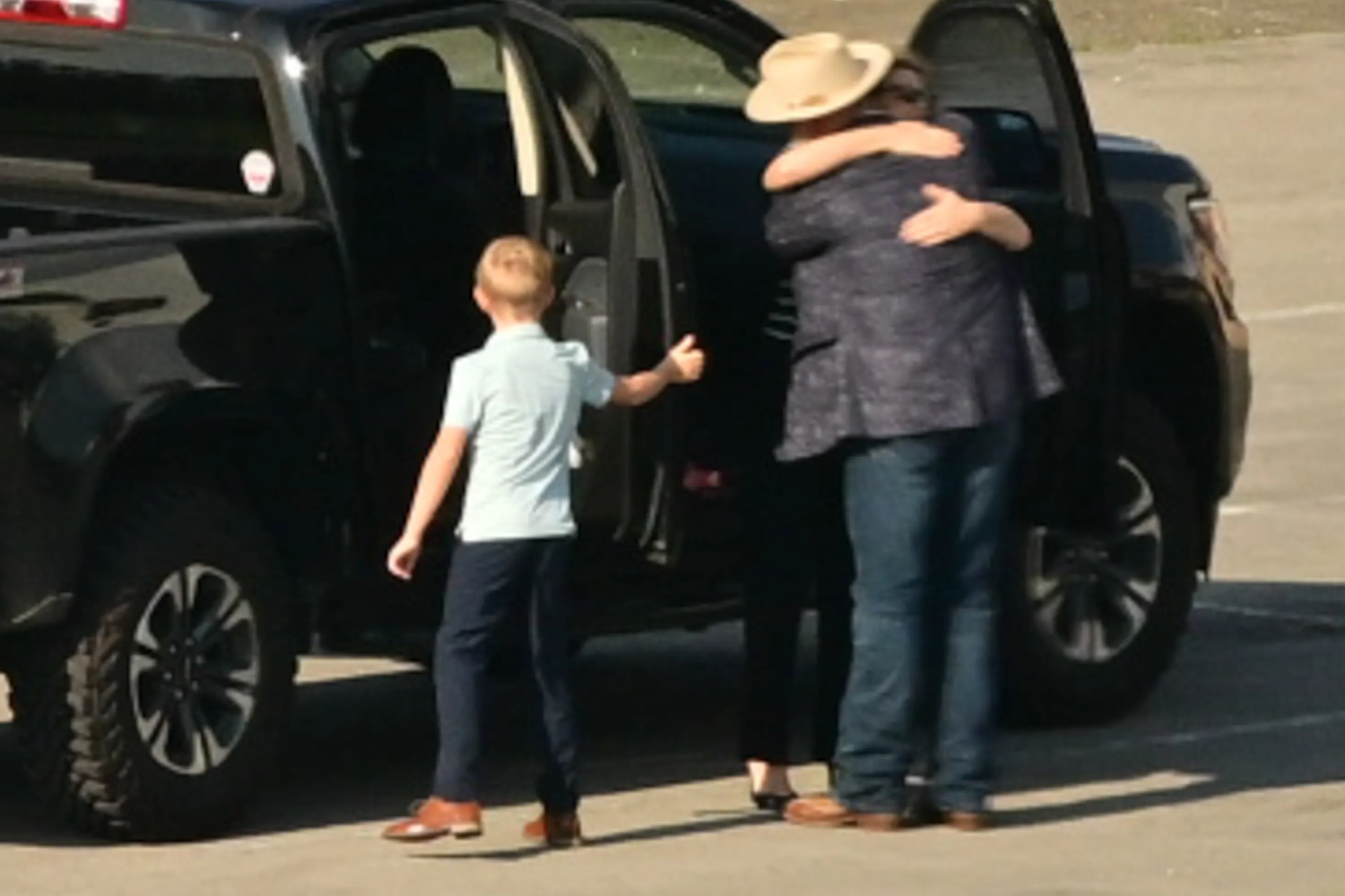 A boy stands next to a truck giving a thumbs up as a man and woman embrace.