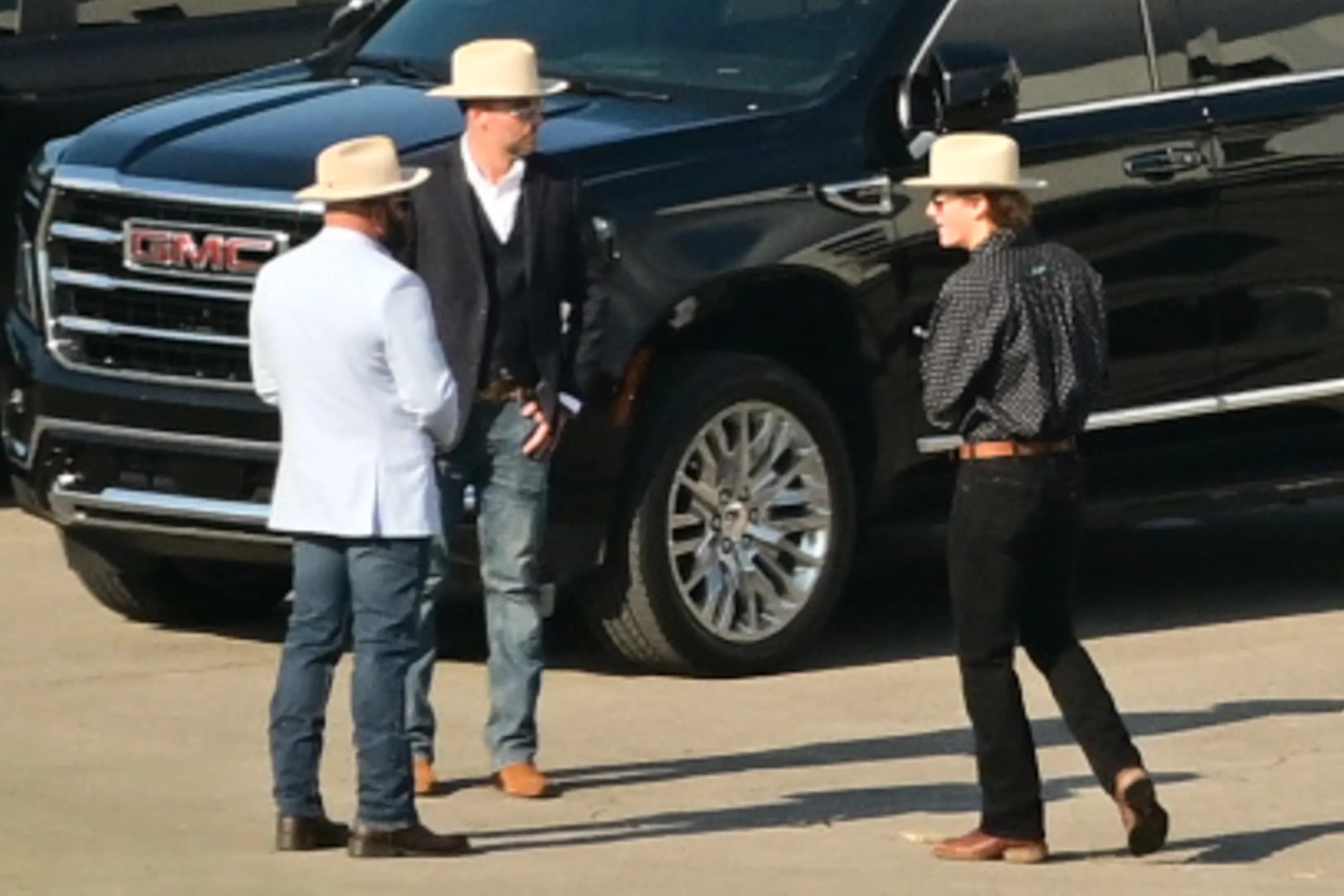Three men in cowboy hats and jeans stand near a black GMC SUV.
