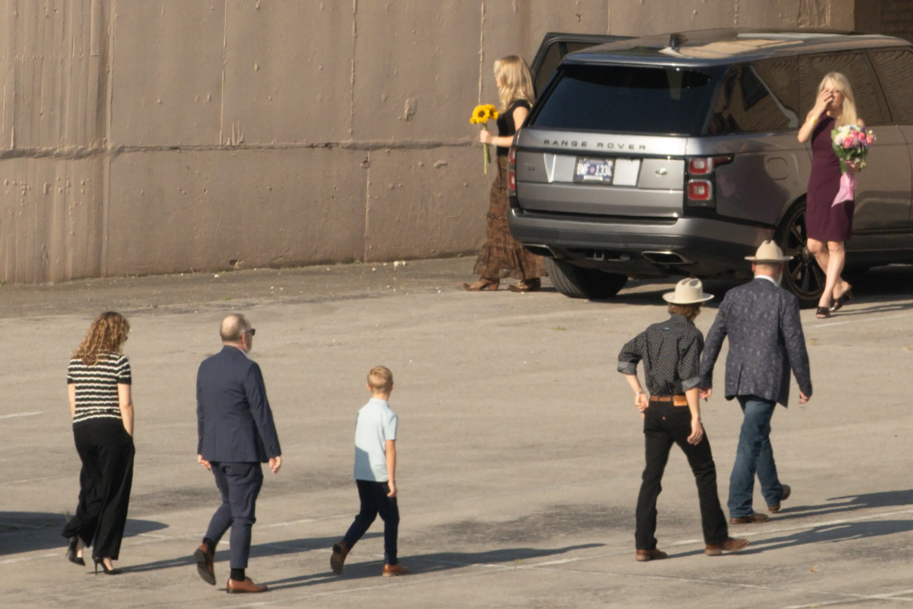 Several people, two carrying flowers, walking across a parking lot away from a Range Rover.