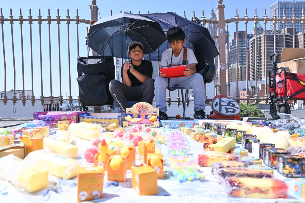 Two boys sell merchandise, including toys and squishies, under black umbrellas on the Brooklyn Bridge.
