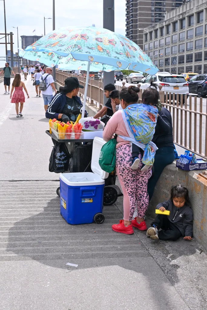 Illegal vendors, including a woman selling fruit cups under an umbrella and a child playing on the ground, on the Brooklyn Bridge pedestrian walkway.