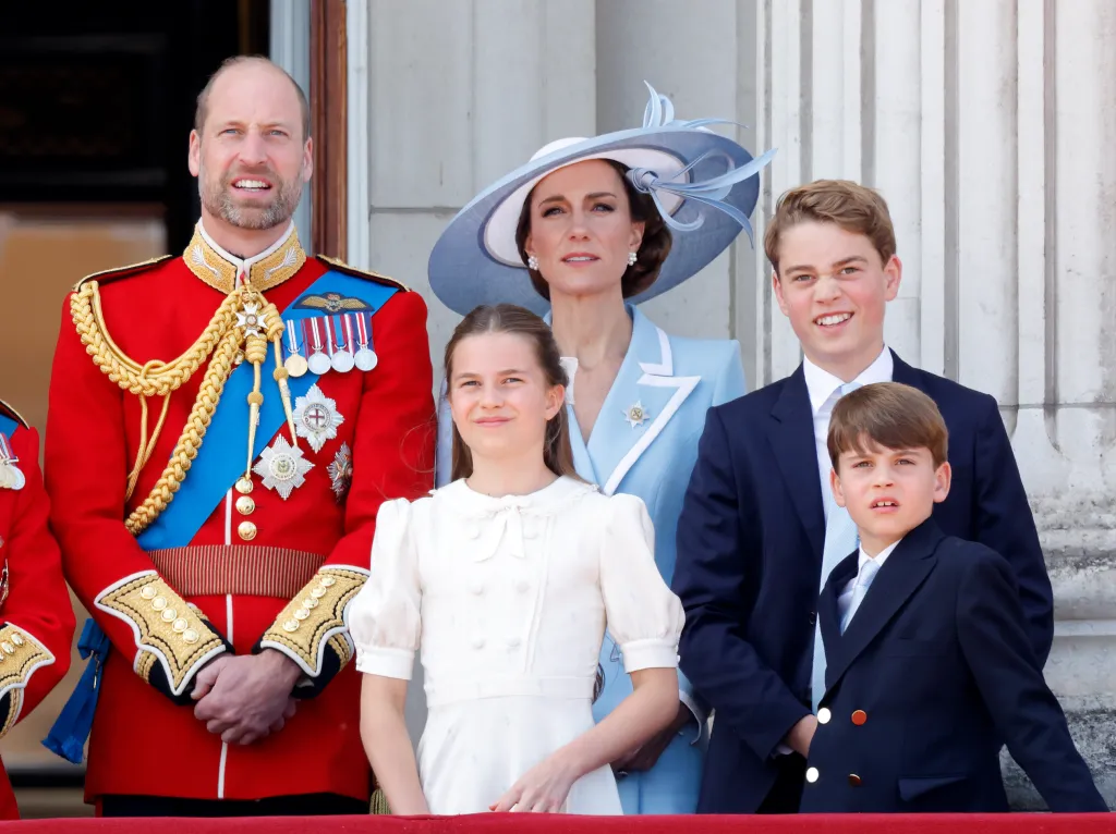 Prince William, in a red military jacket, with medals and blue sash, Princess Catherine, in a blue coat with white trim and blue and white hat, Princess Charlotte, in a white dress, Prince George, in a blue suit, and Prince Louis, in a blue suit, watching an RAF flypast from the balcony of Buckingham Palace after attending Trooping The Colour.