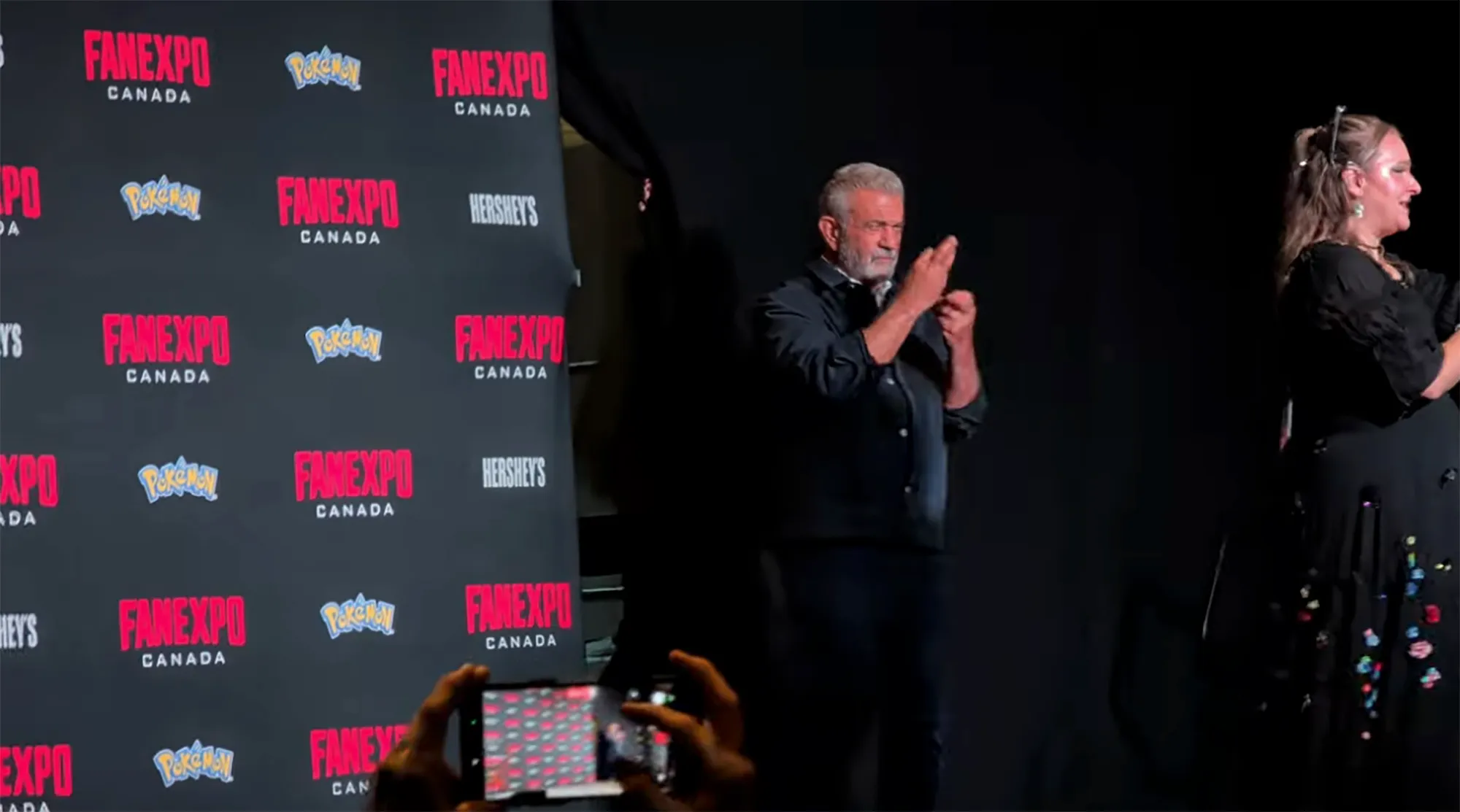 Mel Gibson speaking on stage, making hand gestures as an ASL interpreter stands beside him.