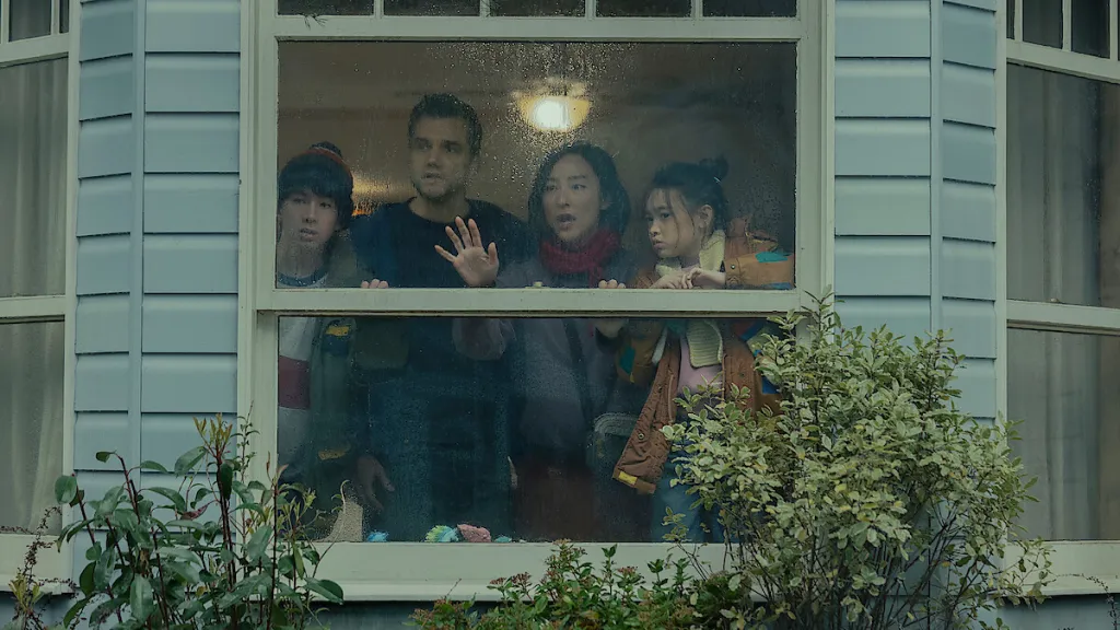 Four people, two adults and two children, look out of a rain-streaked window with expressions of concern.