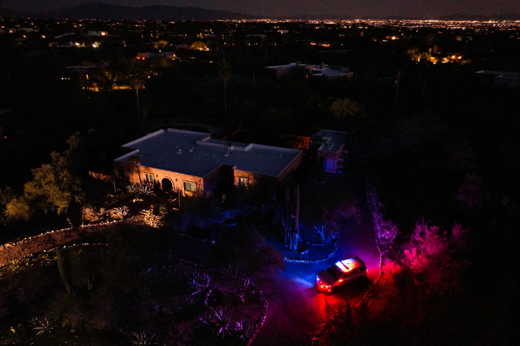 An aerial view at night of Nancy Guthrie's home with a police vehicle with flashing lights on the driveway.