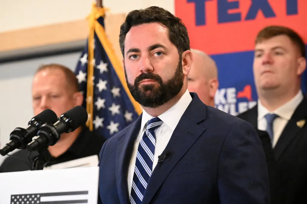 US Representative Mike Lawler at a podium with two microphones, with other men and an American flag behind him.