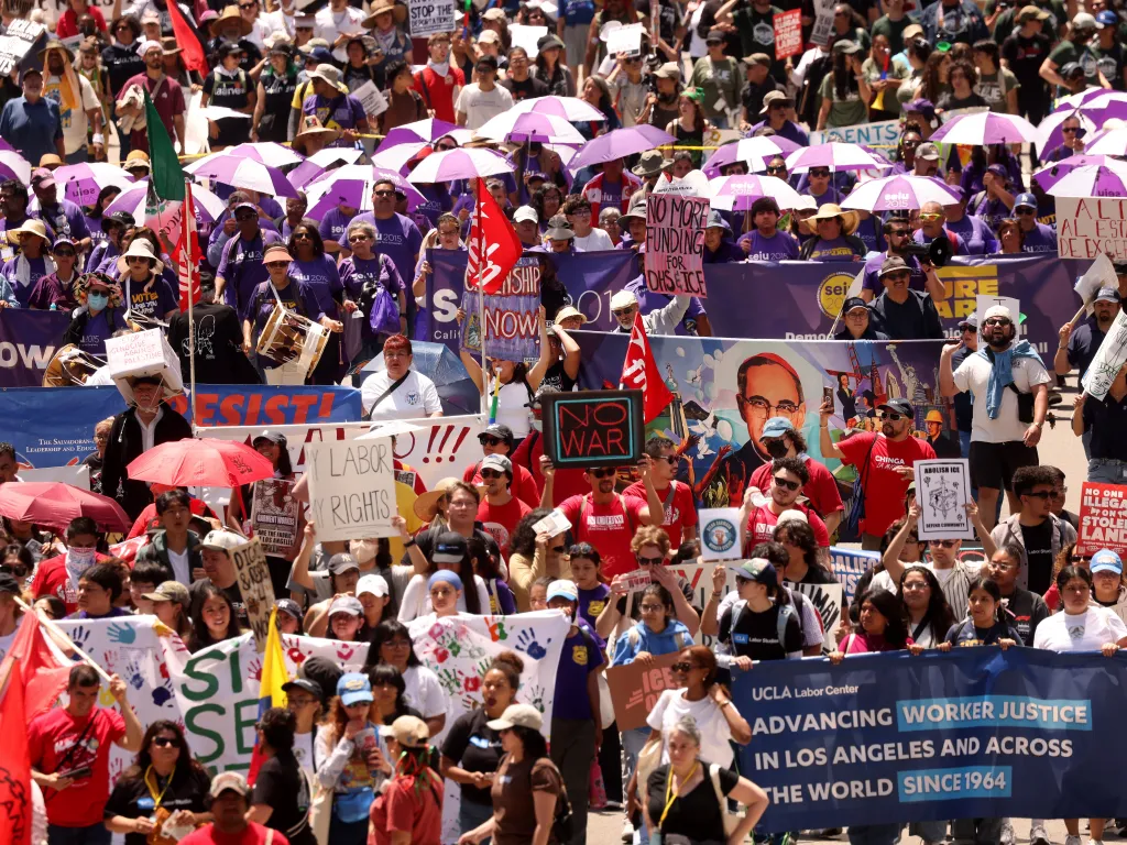 Thousands of workers and activists march in Los Angeles for May Day, many holding signs and banners about worker justice, immigration, and anti-war sentiments, with some holding purple umbrellas.