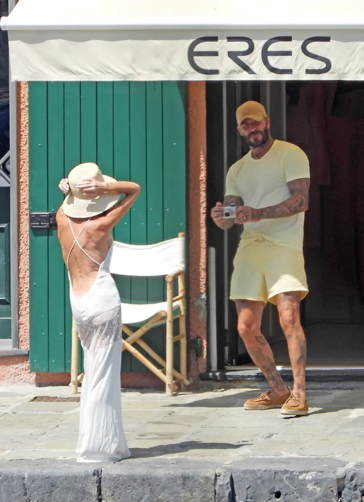 Victoria Beckham posing in a white backless dress and straw hat as David Beckham takes her picture with a vintage digital camera.