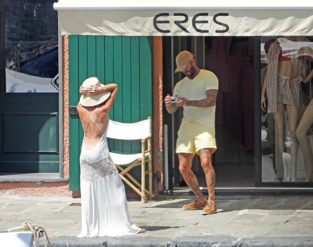 Victoria Beckham poses in a white backless dress and straw hat as David Beckham takes her picture with a vintage digital camera.